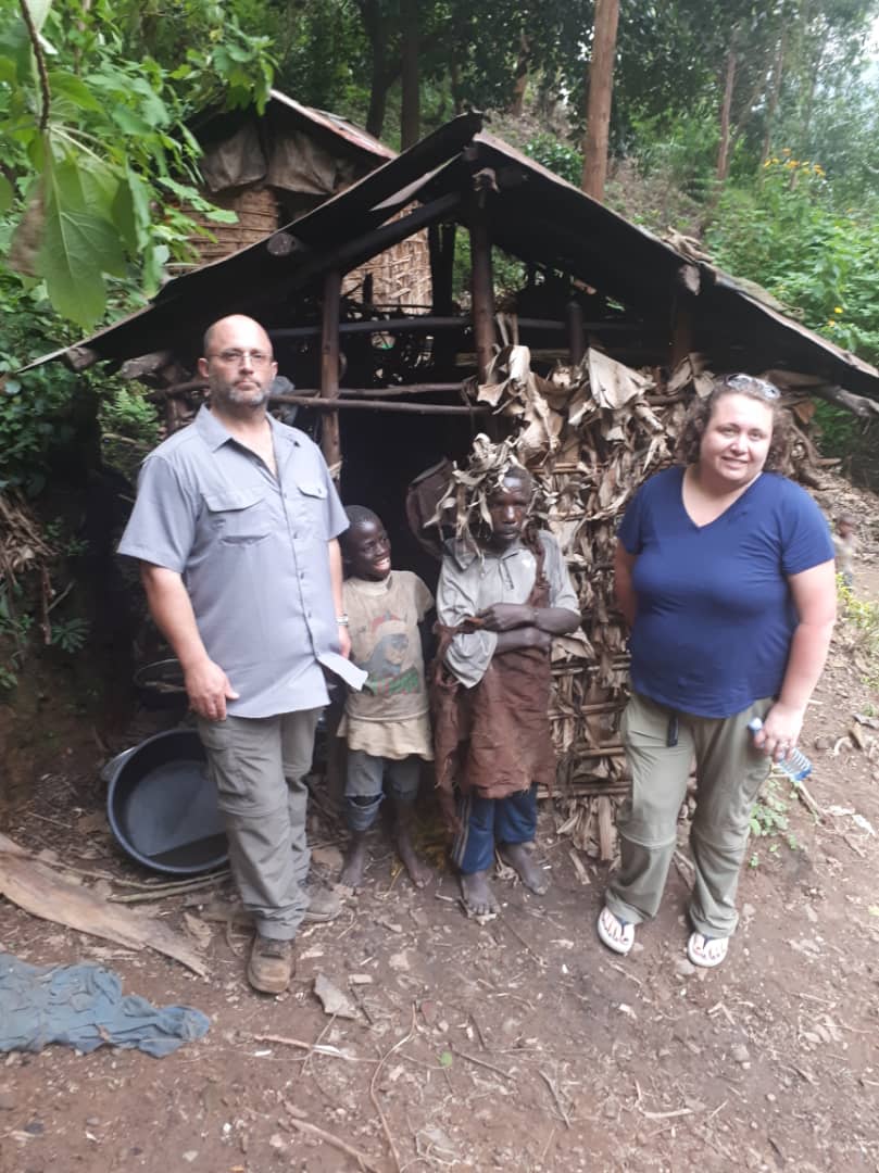 Visitors stand next to a group of Batwa children outside their traditional homestead made of natural materials, showcasing the lifestyle and heritage of the Batwa Pygmy community in Uganda.